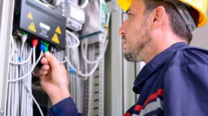 Técnico con casco revisando conexiones en un panel eléctrico con múltiples cables y señalización de alto voltaje.